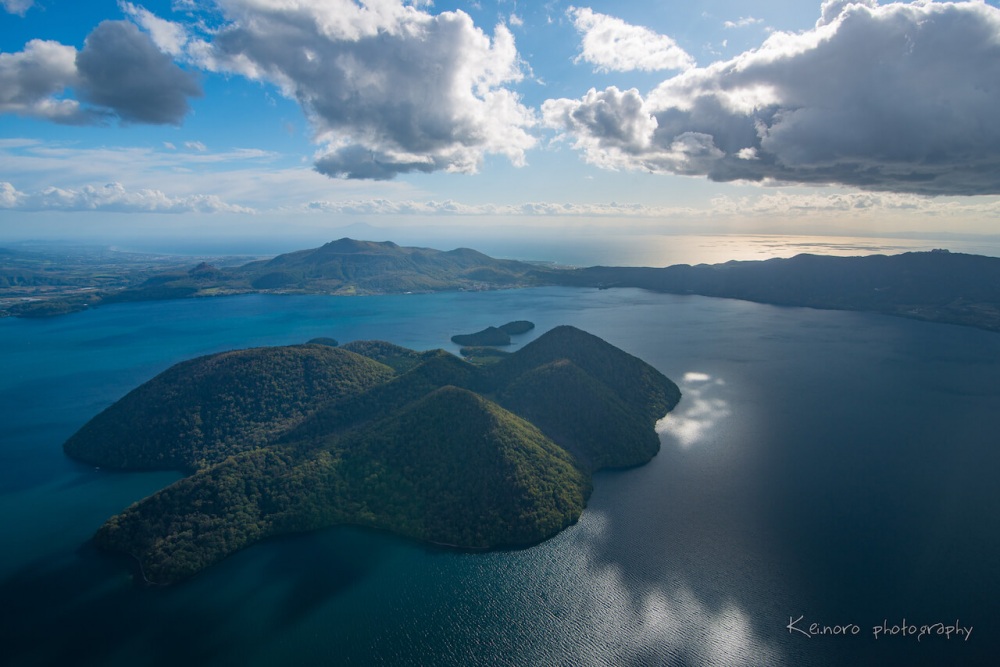 Lake Toya in October, taken from a helicopter in the sky above Nakajima, the island located in the middle of Lake Toya. Visible on the left hand side are Mt. Usu and Mt. Showa Shinzan, which were formed by volcanic eruptions.©Noro Keiichi